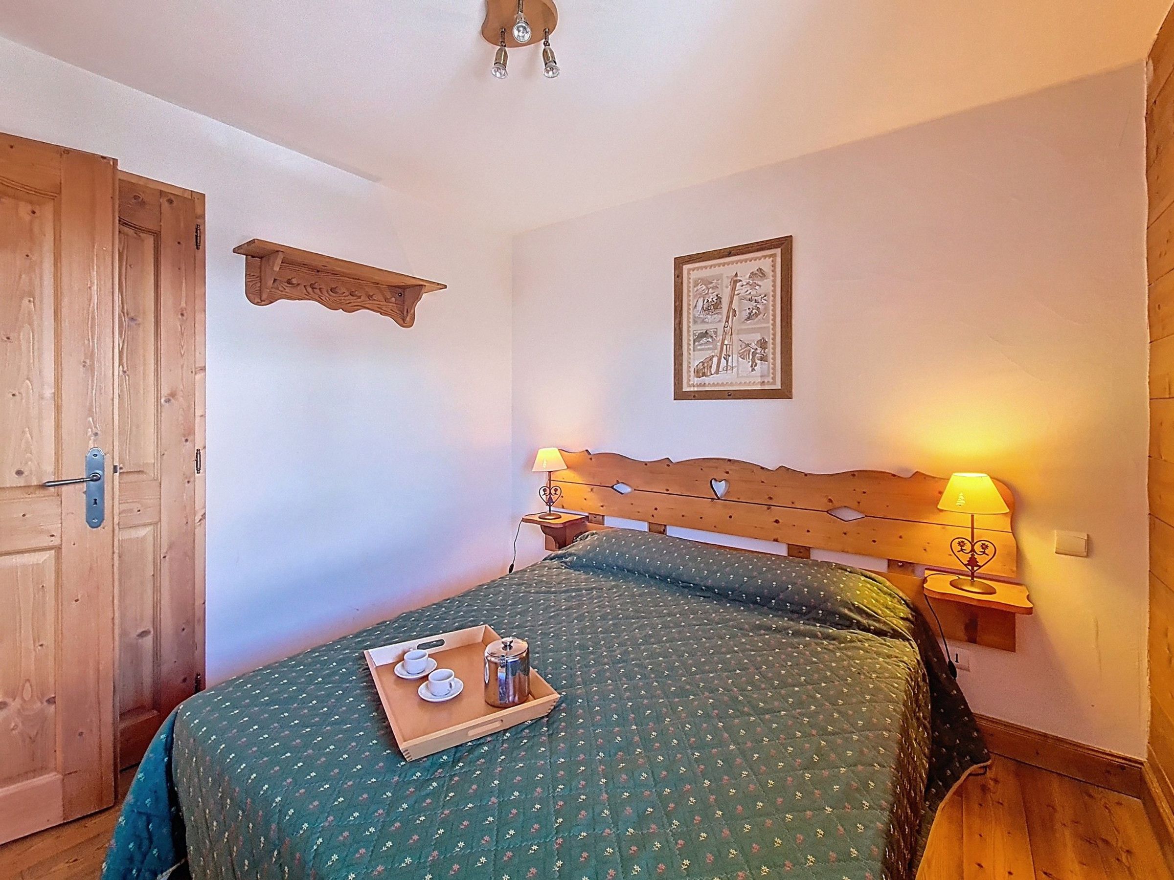 Una habitación con puerta de madera, cama con colcha verde, mesita de noche con lámpara y un cuadro en la pared.