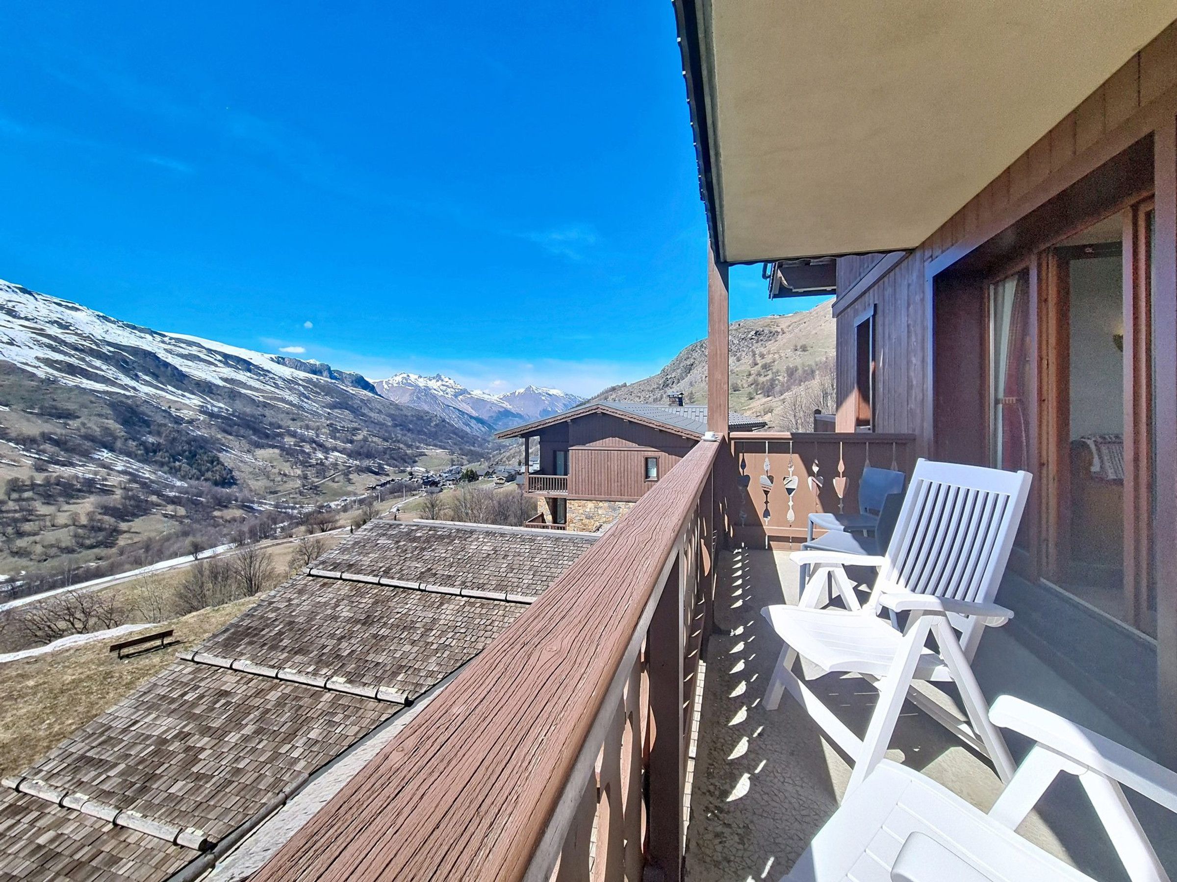 A balcony with a view of snow-capped mountains, a wooden deck, and white chairs.