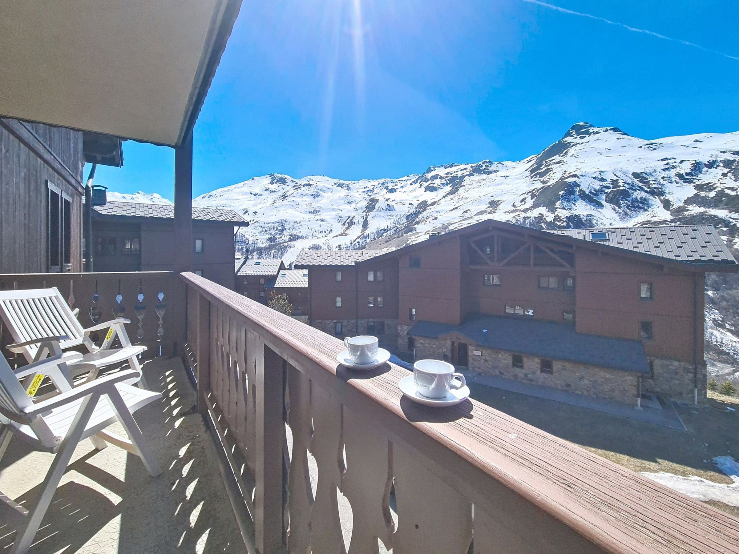 Un balcó amb vistes a muntanyes cobertes de neu, cadires de fusta i una taula amb tasses.