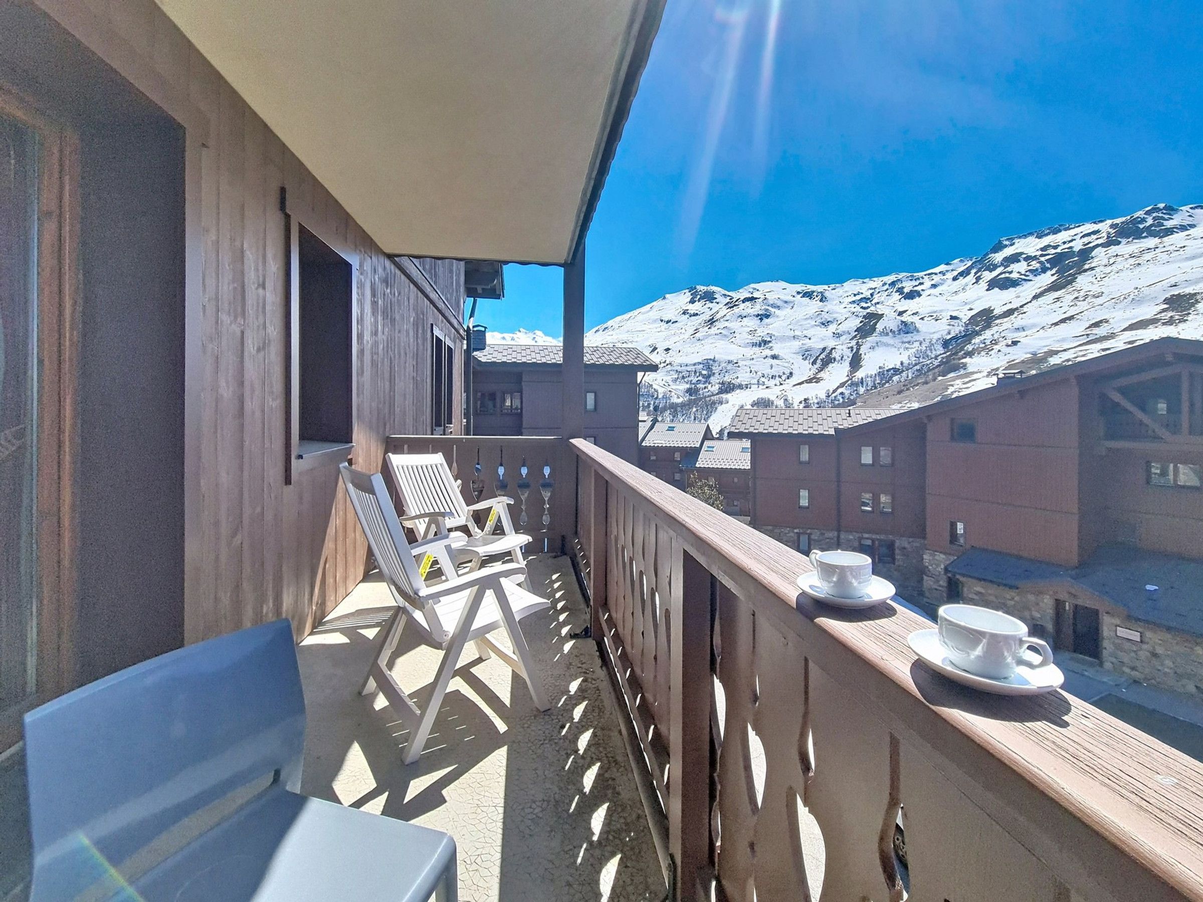 Un balcon avec vue sur des montagnes enneigées, deux chaises blanches et une table avec des tasses.