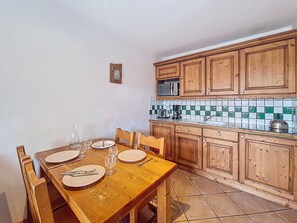 A dining area with a wooden table set for four, a kitchen with wooden cabinets and a tiled backsplash, and a small clock on the wall. - Pleasant apartment - 4 guests (Les Menuires)