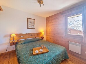 A bedroom with a wooden headboard, a quilted bedspread, bedside lamps, a framed picture, a window with a view, and a tray with cups and a teapot. - Pleasant apartment - 4 guests (Les Menuires)