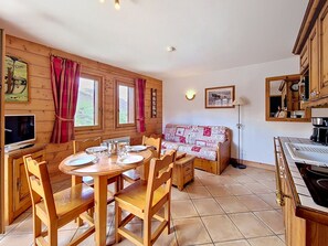 Ein gemütliches Zimmer mit einem Holzessttisch, gedeckt für vier Personen, einer Couch mit gemustertem Bezug und einer Küchenzeile mit Herd und Schränken.