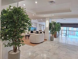 Eine geräumige Lobby mit einer großen Küstertopf-Pflanze, einem gebogenen weißen Sofa und Blick durch eine Glastür.