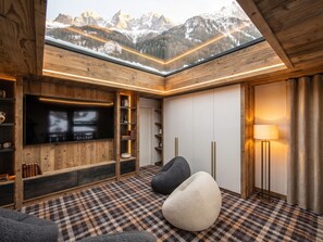 Ein gemütliches Zimmer mit einem großen Fernseher, Holzregalen, einem gemusterten Teppich und Bergblick durch ein Oberlicht.