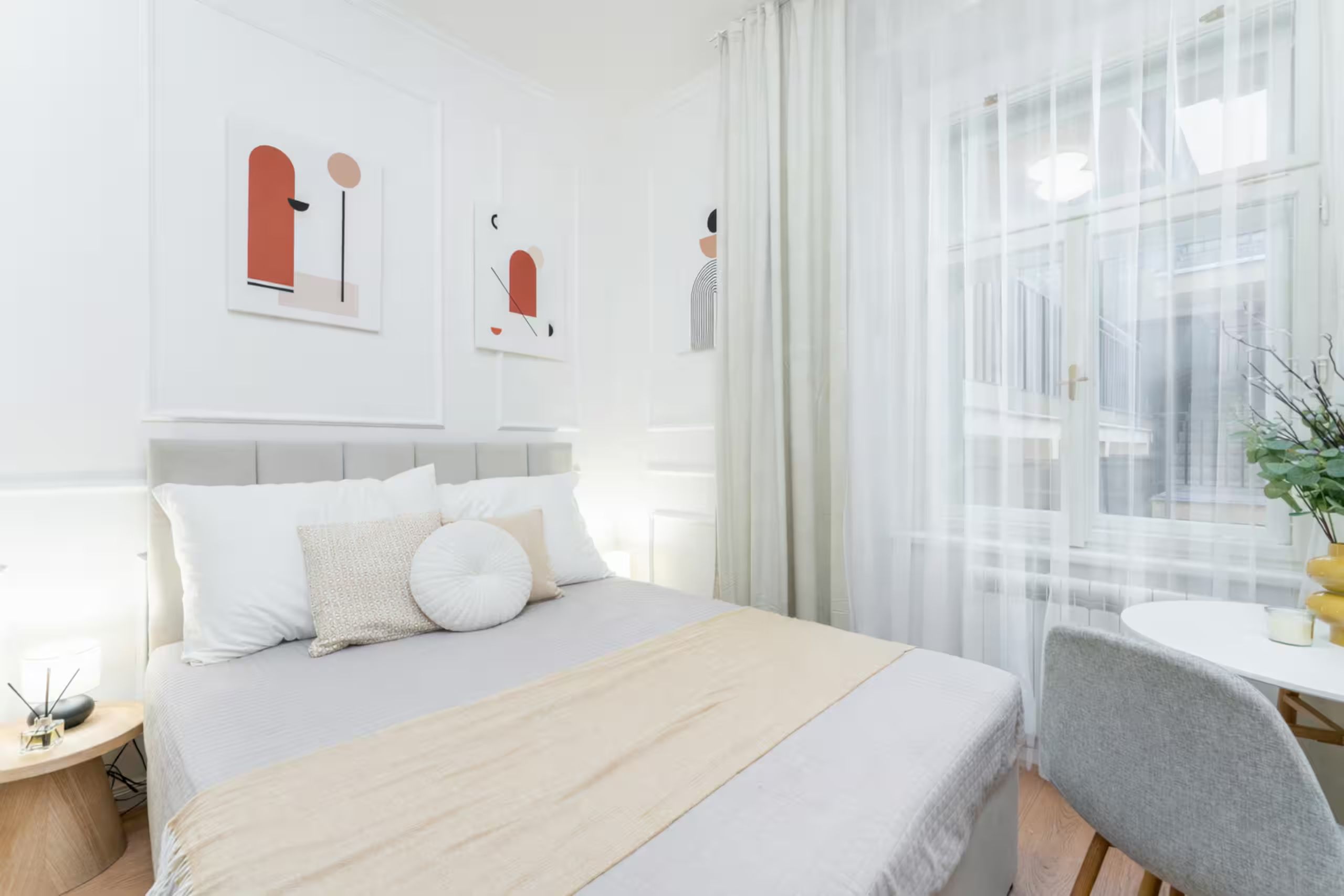 A modern hotel room with a bed, bedside table, and a view of the city through sheer curtains.