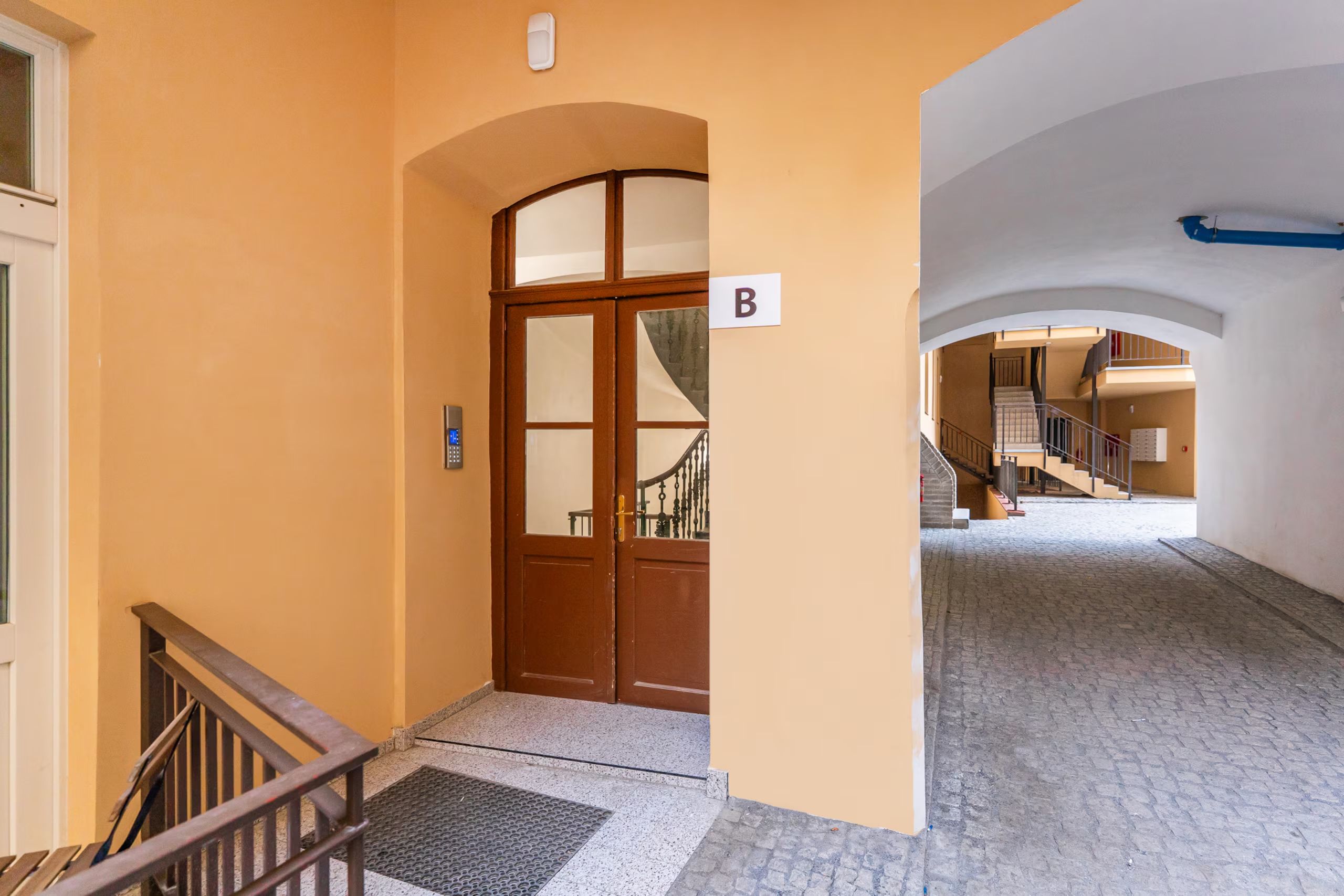 A hallway with a wooden door marked 'B', a metal handrail, and a tiled floor.