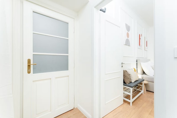 Un couloir avec une porte blanche ornée d’un panneau en verre dépoli, un mur blanc avec une porte, et un lit avec un cadre blanc et un oreiller.
