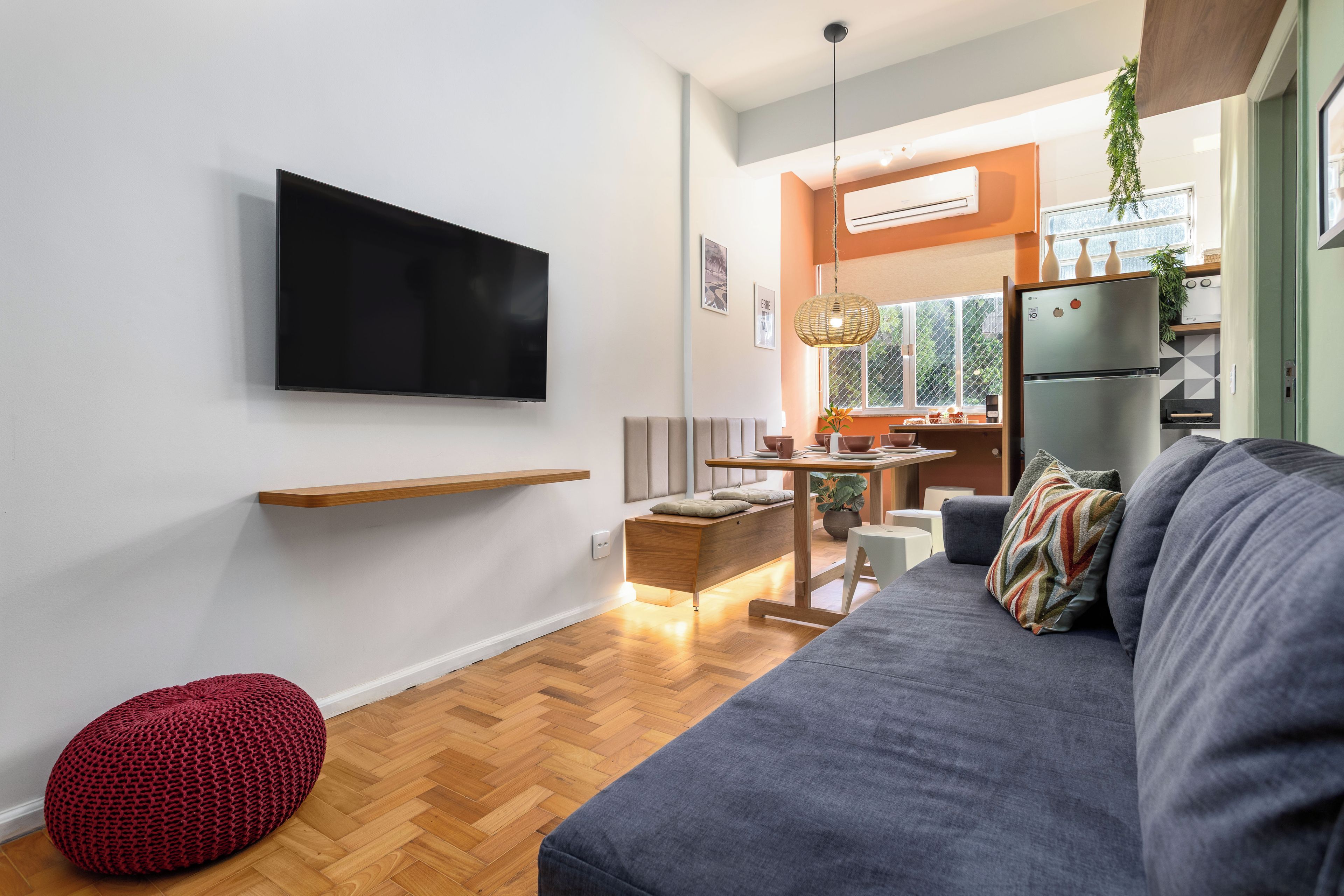 A modern living room with a flat-screen TV, a grey sofa, a red pouf, and a wooden dining table.