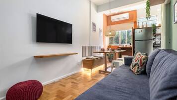 A modern living room with a flat-screen TV, a grey sofa, a red pouf, and a wooden dining table.