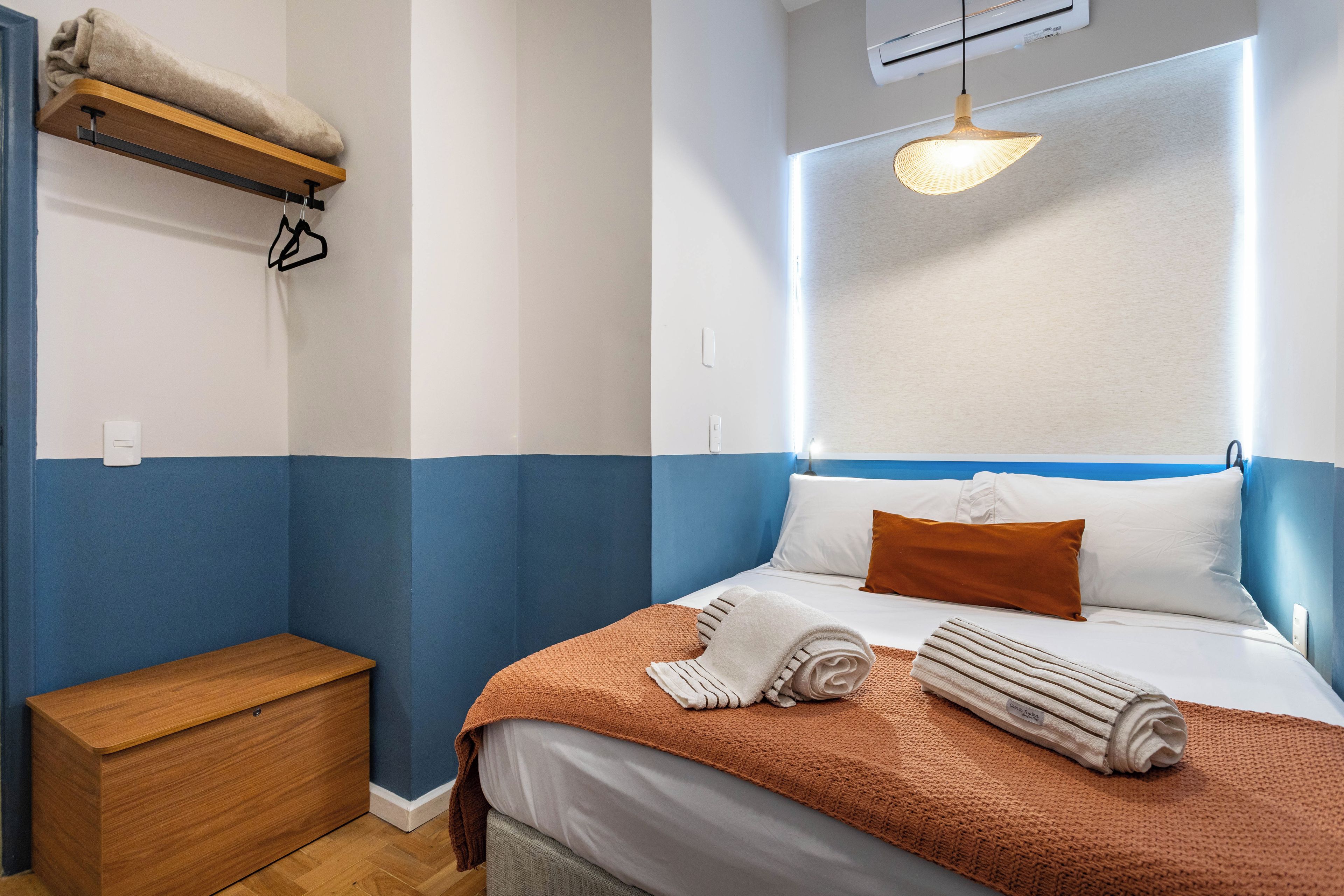 Une chambre d’hôtel comprenant un lit, une table de chevet, un petit banc en bois, un luminaire suspendu et un appareil de climatisation fixé au mur.