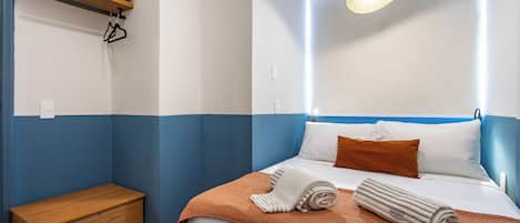 Une chambre d’hôtel comprenant un lit, une table de chevet, un petit banc en bois, un luminaire suspendu et un appareil de climatisation fixé au mur.
