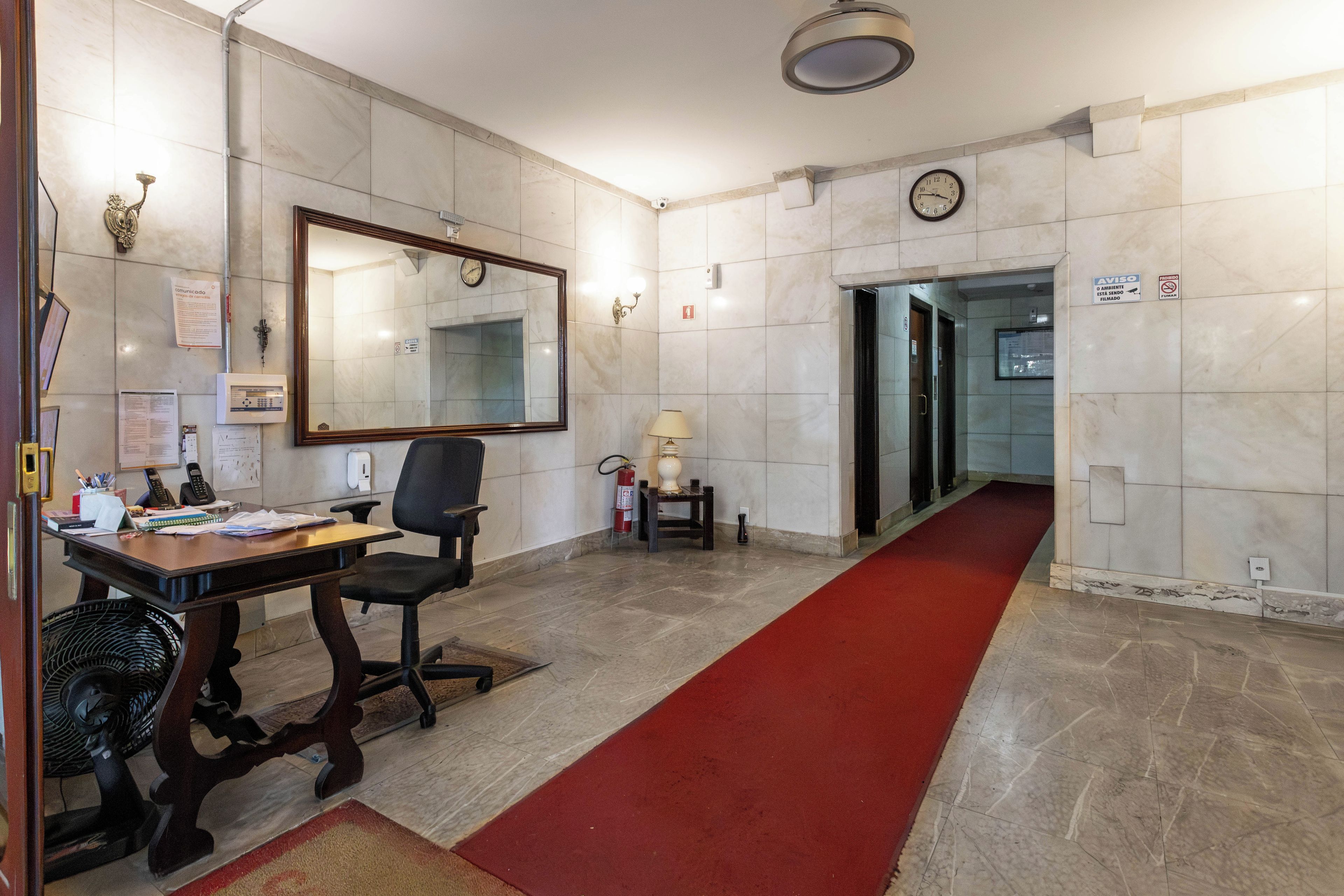 Un hall d’hôtel avec un comptoir d’accueil, un grand miroir, une horloge et un tapis rouge menant à un ascenseur.