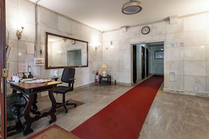 A hotel lobby with a reception desk, a large mirror, a clock, and a red carpet leading to an elevator.