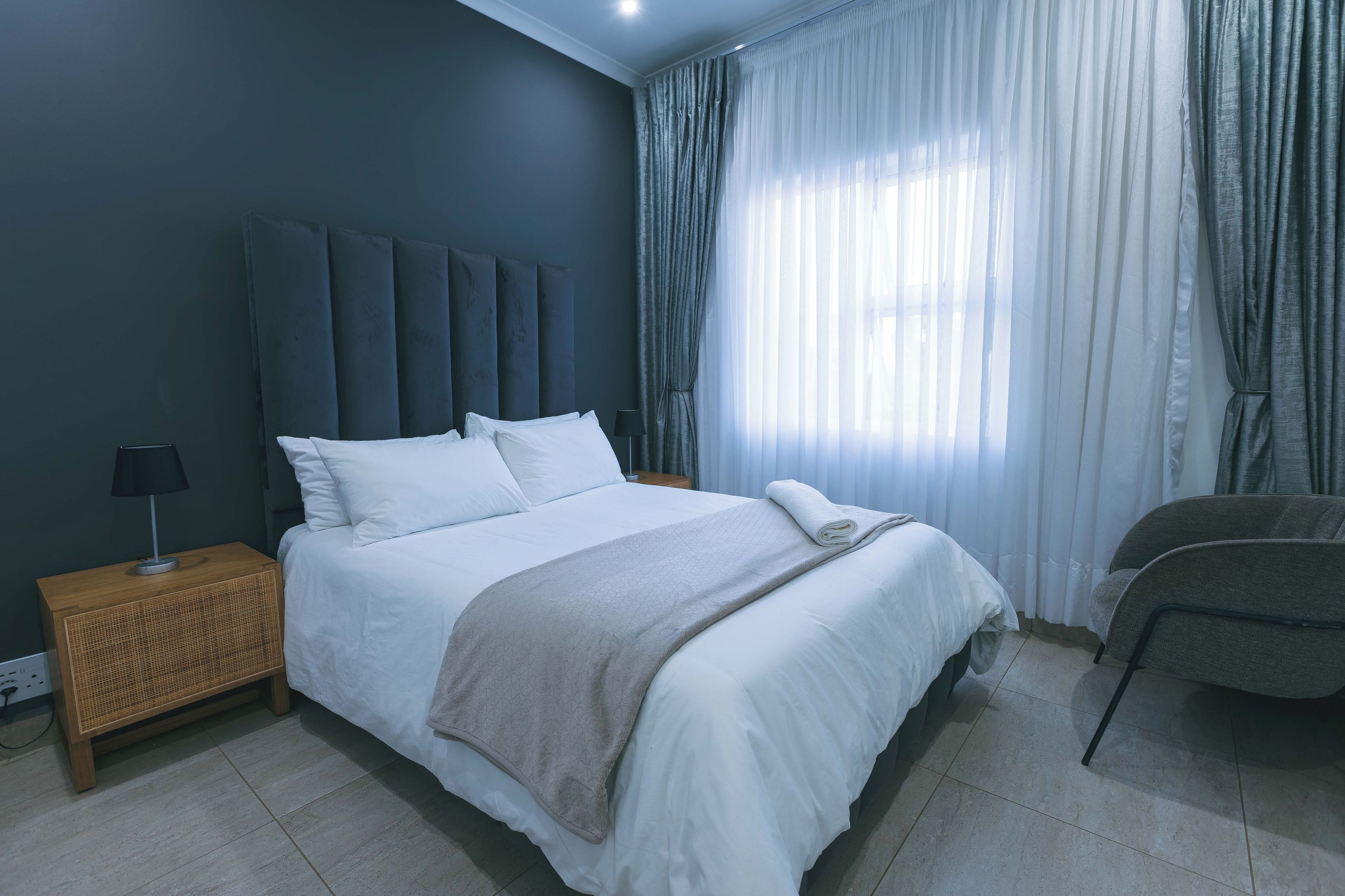 Un dormitorio con cama, mesita de noche, lámpara, una silla y ventana con cortinas.