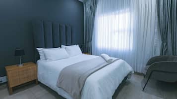 Un dormitorio con cama, mesita de noche, lámpara, una silla y ventana con cortinas.