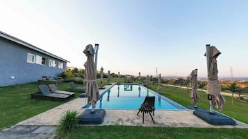 Una piscina con sillones de descanso y sombrillas, rodeada de césped y un edificio.