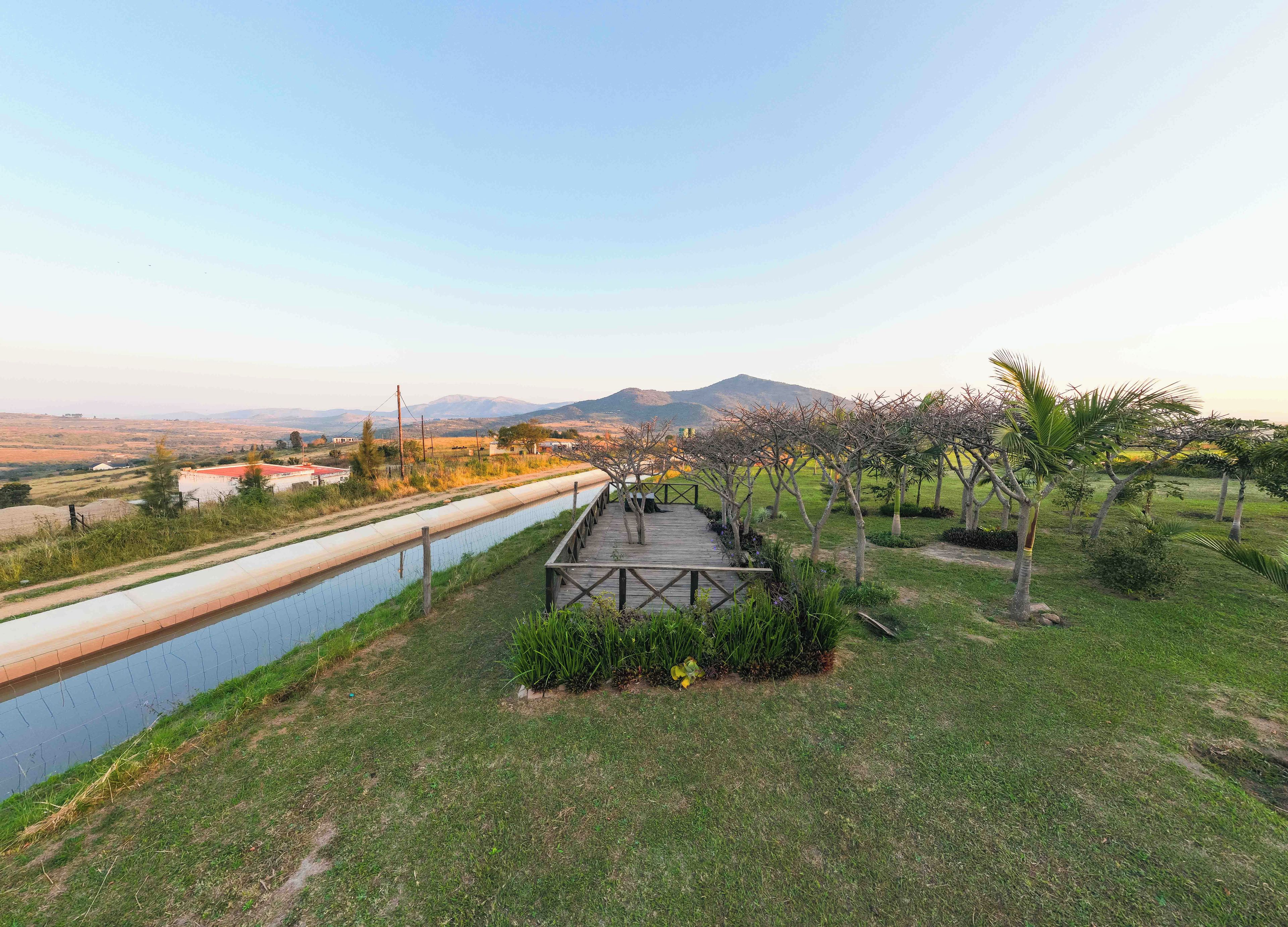 Un camí que condueix a una terrassa de fusta amb vista a una zona paisatgística, un canal d'aigua i muntanyes al lluny.