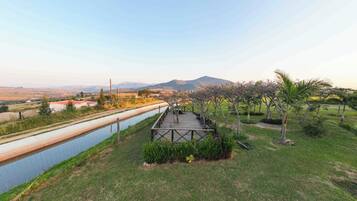 Un sentier mène à une terrasse en bois qui surplombe un espace aménagé avec un canal d’eau et des montagnes au loin.