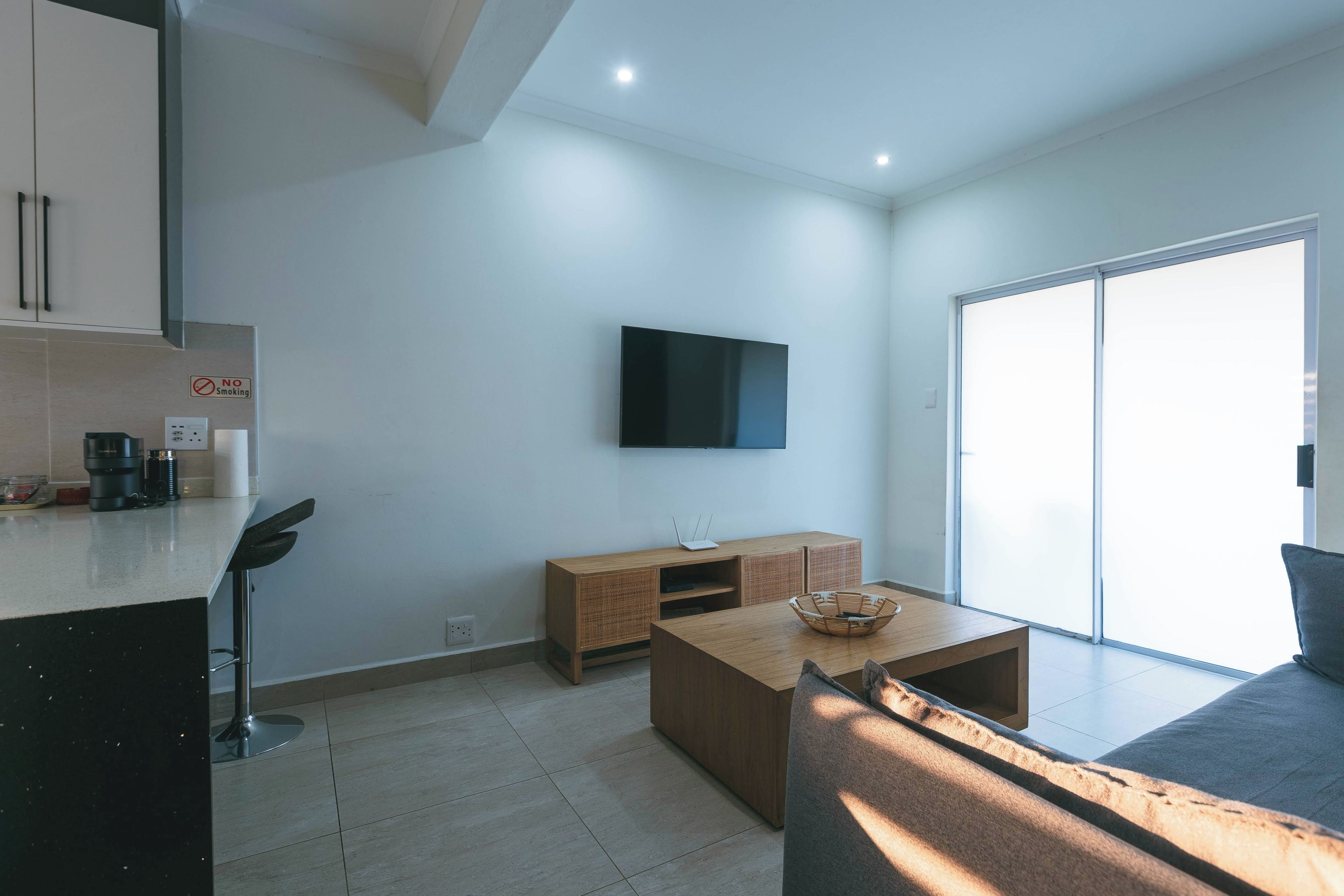 Un espacio de estar moderno con un televisor de pantalla plana en la pared, una mesa de centro de madera, un sofá y una zona de cocina con una cafetera.