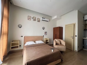 A hotel room with a bed, a chair, a wardrobe, a desk, and an air conditioning unit.