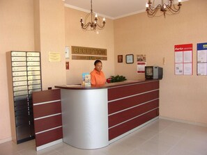 Un mostrador de recepción con una mujer detrás, una araña, un reloj y un tablón de anuncios con avisos.