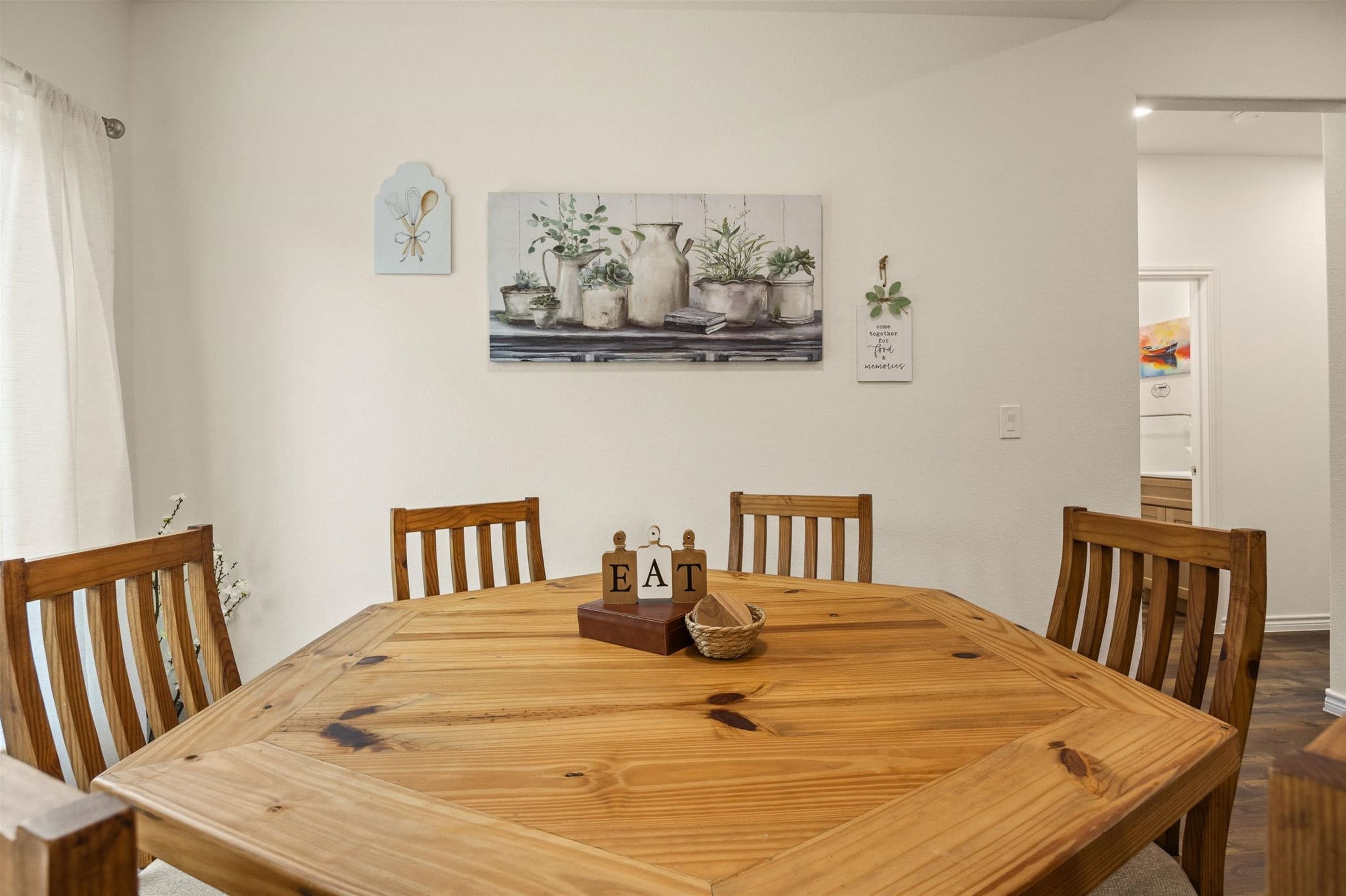 A dining area with a wooden table, four chairs, and a painting of potted plants on the wall.