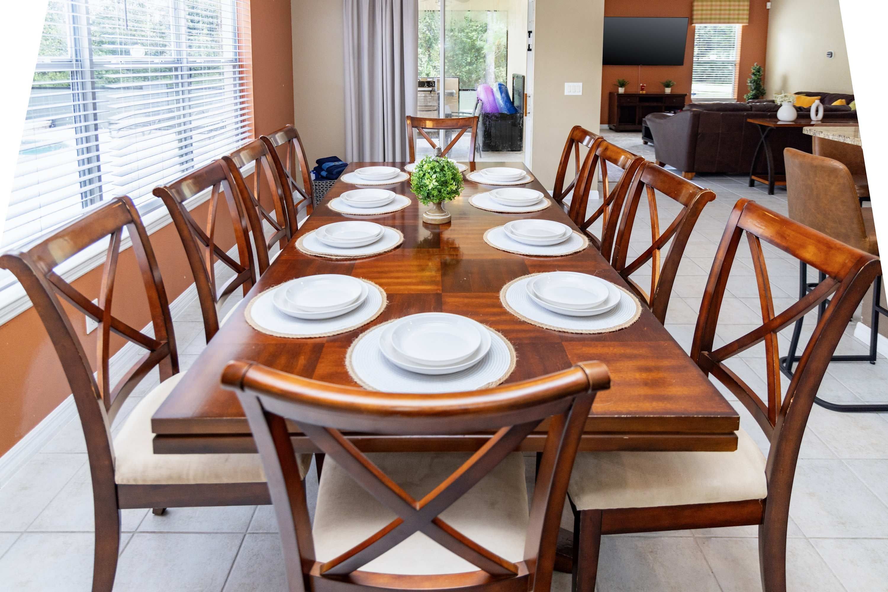 Uma sala de jantar com uma longa mesa de madeira posta para refeição, rodeada por cadeiras de madeira com assentos acolchoados.