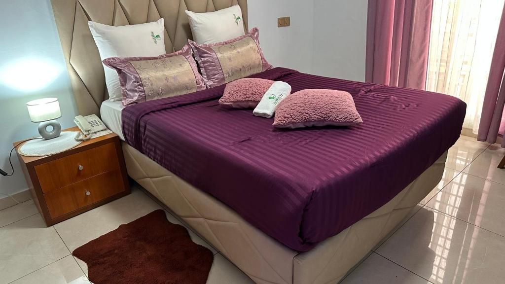 A neatly made bed with a purple bedspread and matching pillows, a wooden nightstand with a lamp and phone, and a brown rug on the floor.