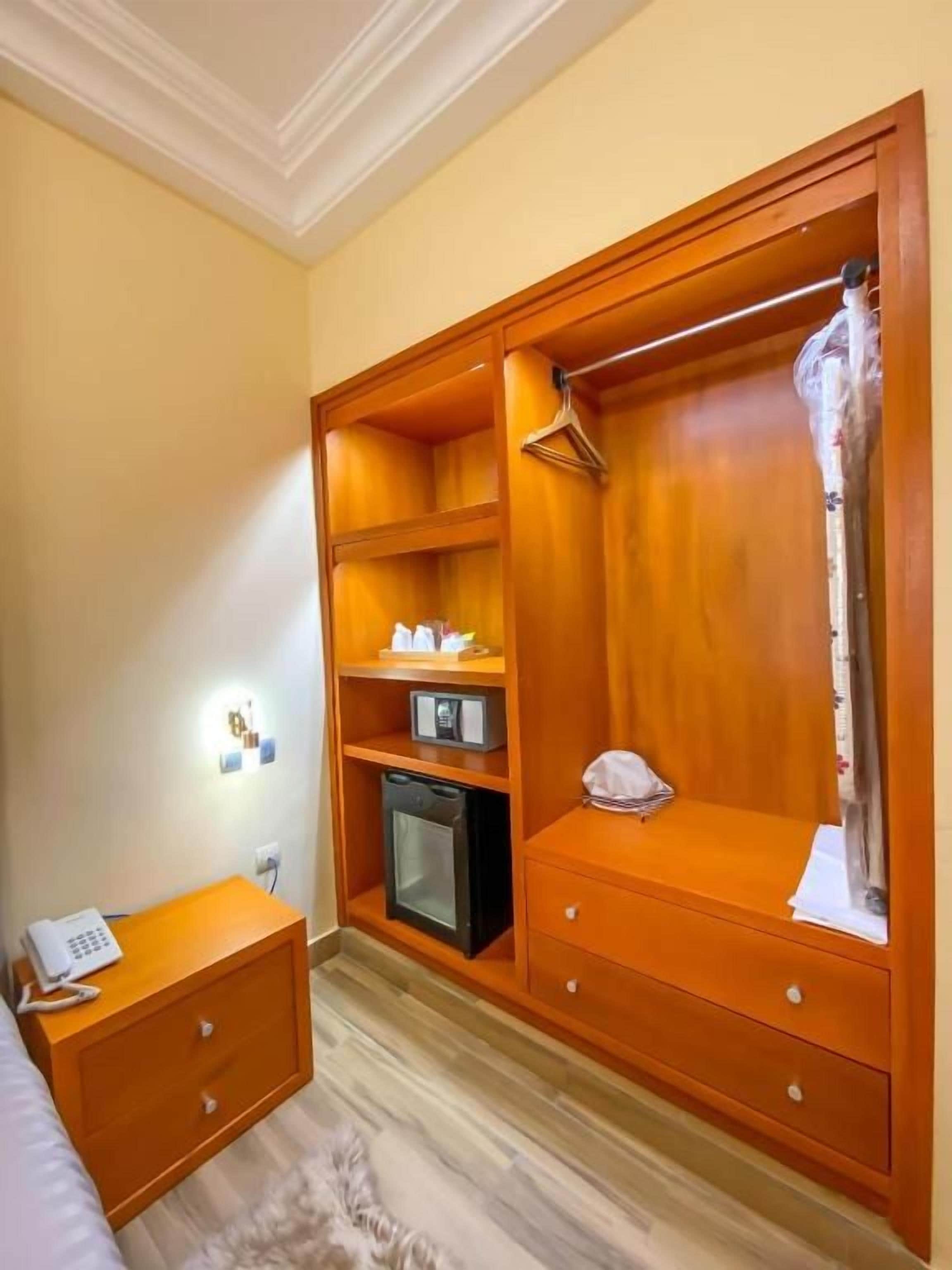 A bedroom with a wooden wardrobe, a bedside table with a phone, a microwave, and a small cabinet.