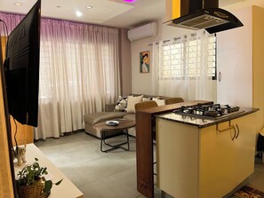 Une cuisine moderne équipée d’une cuisinière à gaz intégrée, un coin repas avec un canapé et une petite table, et un salon avec une grande télévision à écran plat et un tableau au mur.
