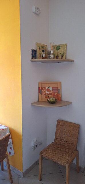 A room with a yellow wall, a wicker chair, two wooden shelves with decorative items, and a power outlet.