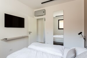 Un dormitorio moderno con una cama, un televisor montado, un estante y una puerta que da a otra habitación.