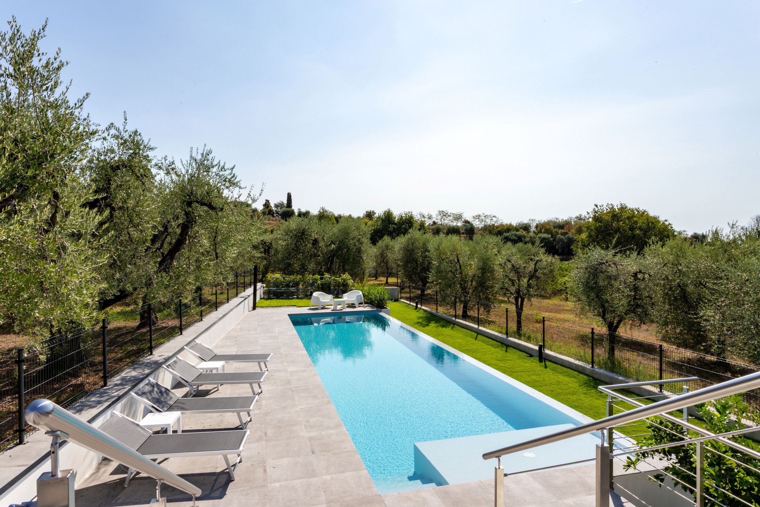 Une piscine rectangulaire à l’eau d’un bleu limpide, entourée d’un petit muret en pierre. Des chaises longues blanches sont alignées le long du bord de la piscine, et un escalier en métal avec une rampe descend vers la piscine. L’ensemble est délimité par une clôture en métal noir, et au-delà de la clôture, on trouve une variété de verdure, notamment des arbres et des arbustes.