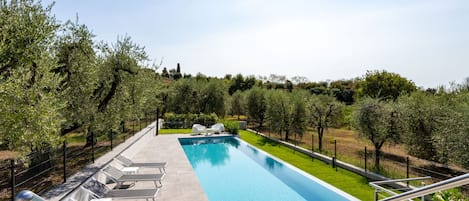 Une piscine rectangulaire à l’eau d’un bleu limpide, entourée d’un petit muret en pierre. Des chaises longues blanches sont alignées le long du bord de la piscine, et un escalier en métal avec une rampe descend vers la piscine. L’ensemble est délimité par une clôture en métal noir, et au-delà de la clôture, on trouve une variété de verdure, notamment des arbres et des arbustes.