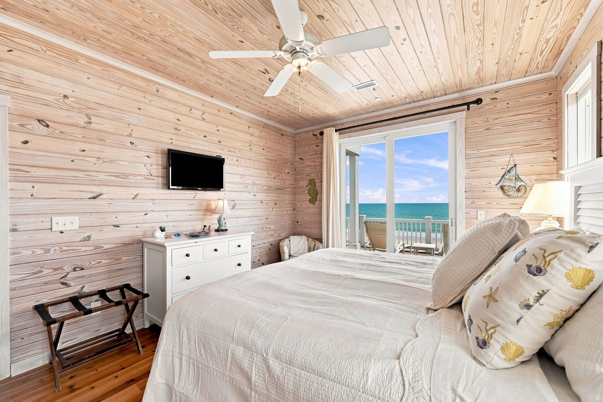 Ein Schlafzimmer mit Holzbalkendecke, einem Bett mit weißer Bettdecke, einem Fernseher an der Wand und Meerblick durch eine Schiebetür.
