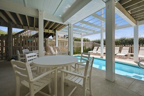 Un espacio exterior techado con piscina, mobiliario blanco y una valla de madera.