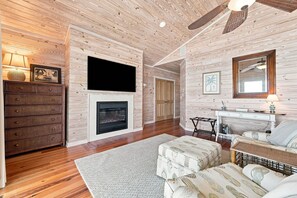 A living room with a fireplace, wooden floors, a TV mounted on the wall, and a sofa. - The Beach House #226 - Luxury 8-Bedroom Oceanfront Rental w/ Private Pool (North Topsail Beach)