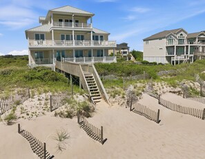 A beachfront property with multiple-story buildings, a wooden staircase leading to a deck, and a sandy beach area with a fence. - The Beach House #226 - Luxury 8-Bedroom Oceanfront Rental w/ Private Pool (North Topsail Beach)
