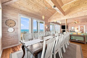 Un comedor amplio con una larga mesa de madera y sillas blancas, ventanas grandes que ofrecen vistas al océano, y una zona de estar acogedora con un sofá y televisor.
