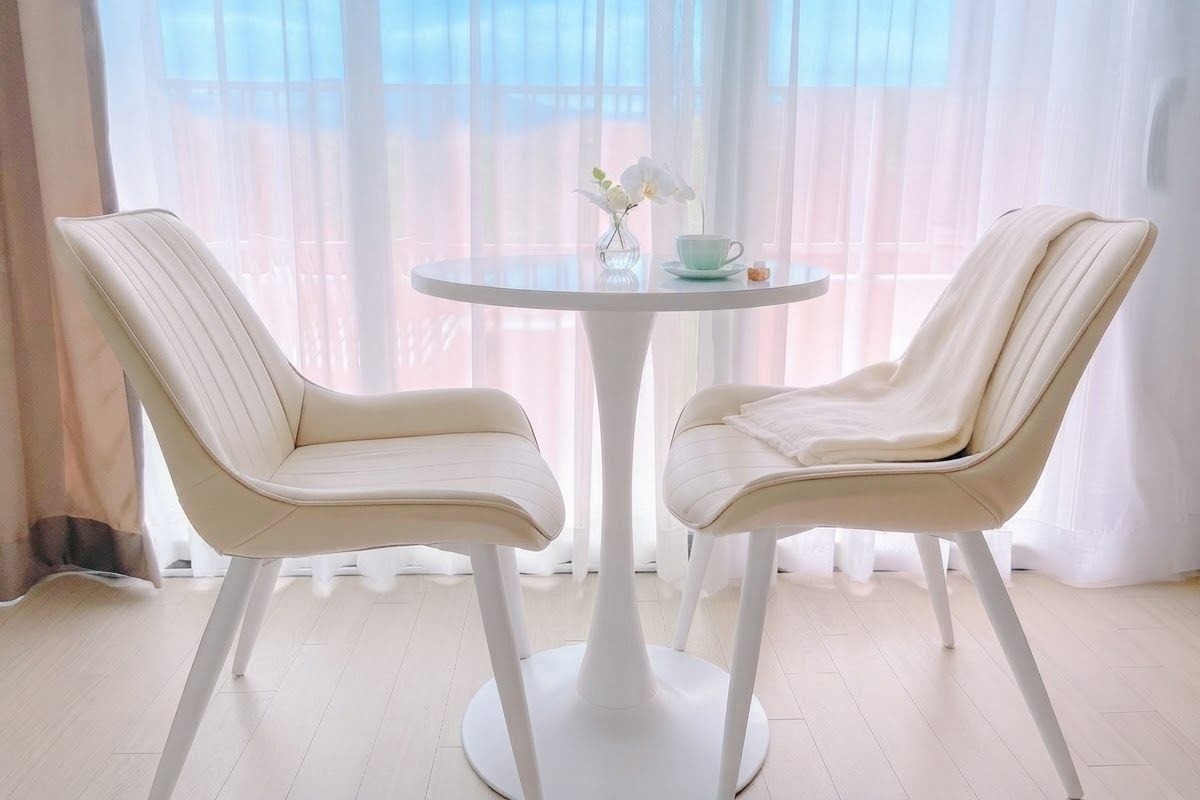 Una habitación con dos sillas blancas alrededor de una pequeña mesa redonda, un jarrón con flores y cortinas traslúcidas.