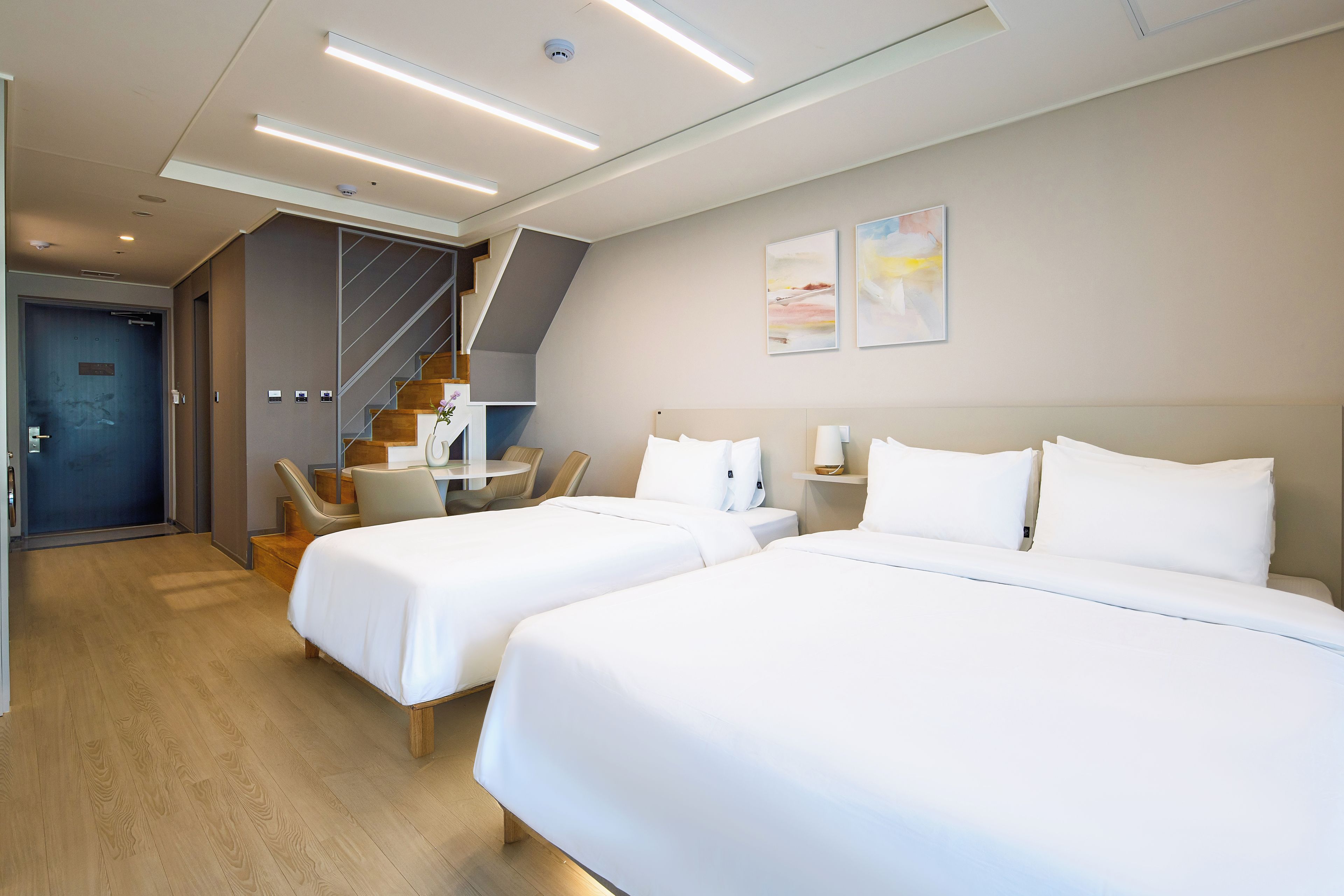 A hotel room with two beds, a desk, a chair, a staircase, and artwork on the wall.