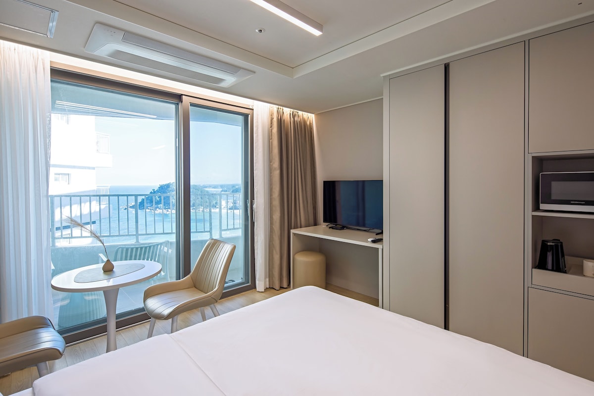 A hotel room with a bed, a TV on a stand, a small table with a chair, and a view of the city through large windows.