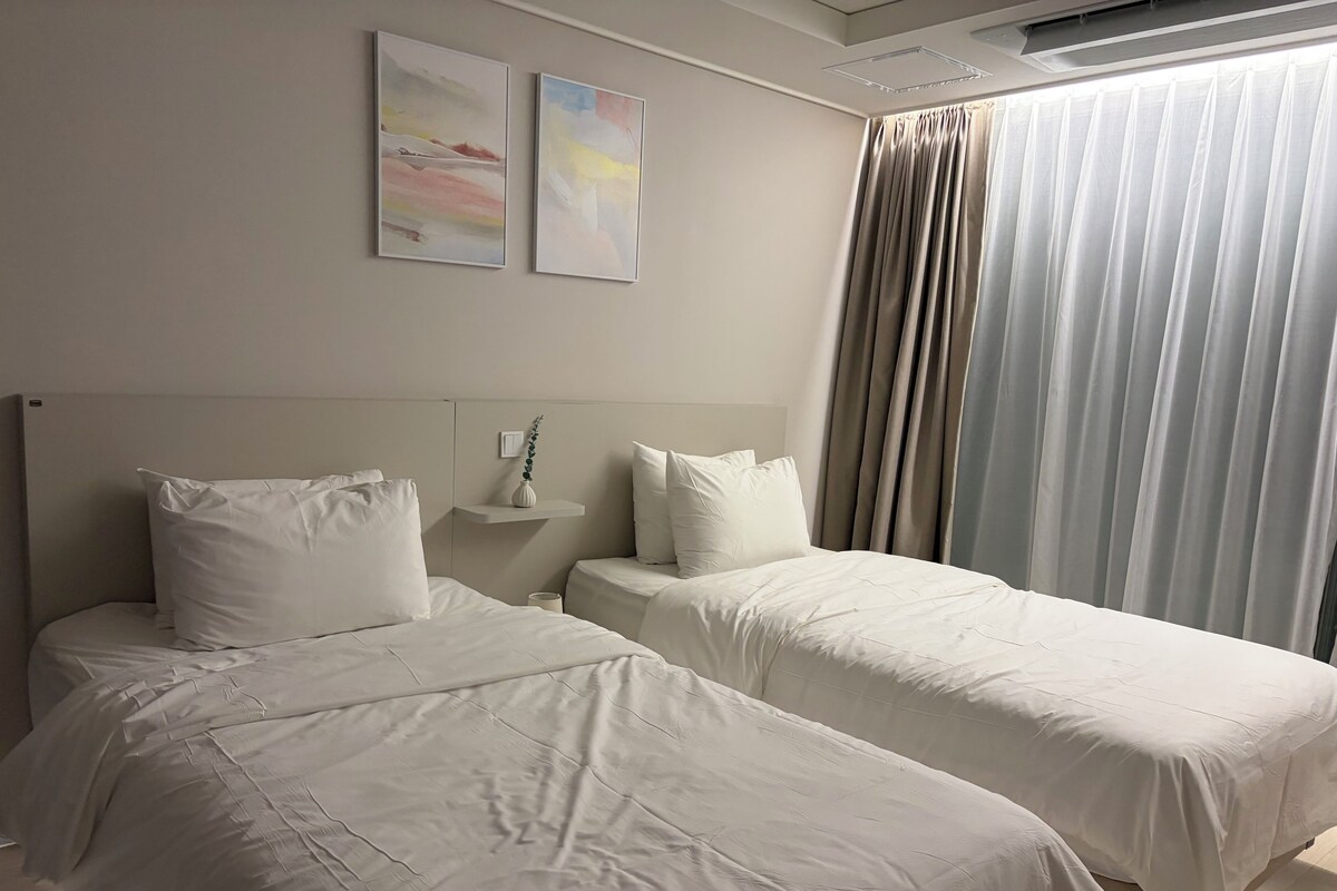 Habitación de hotel con dos camas, una mesita de noche, un interruptor de luz en la pared y una ventana con cortinas.