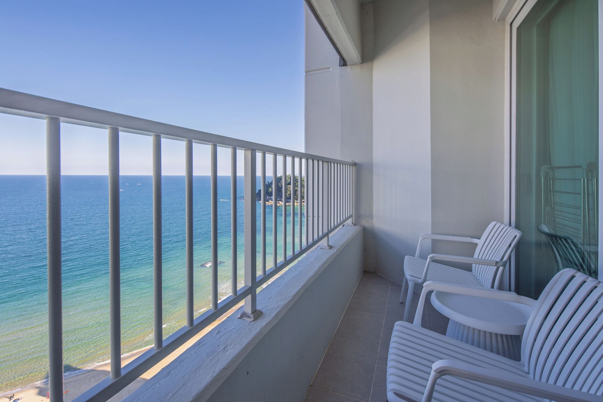 A balcony with a metal railing, white plastic chairs, and a view of the sea.