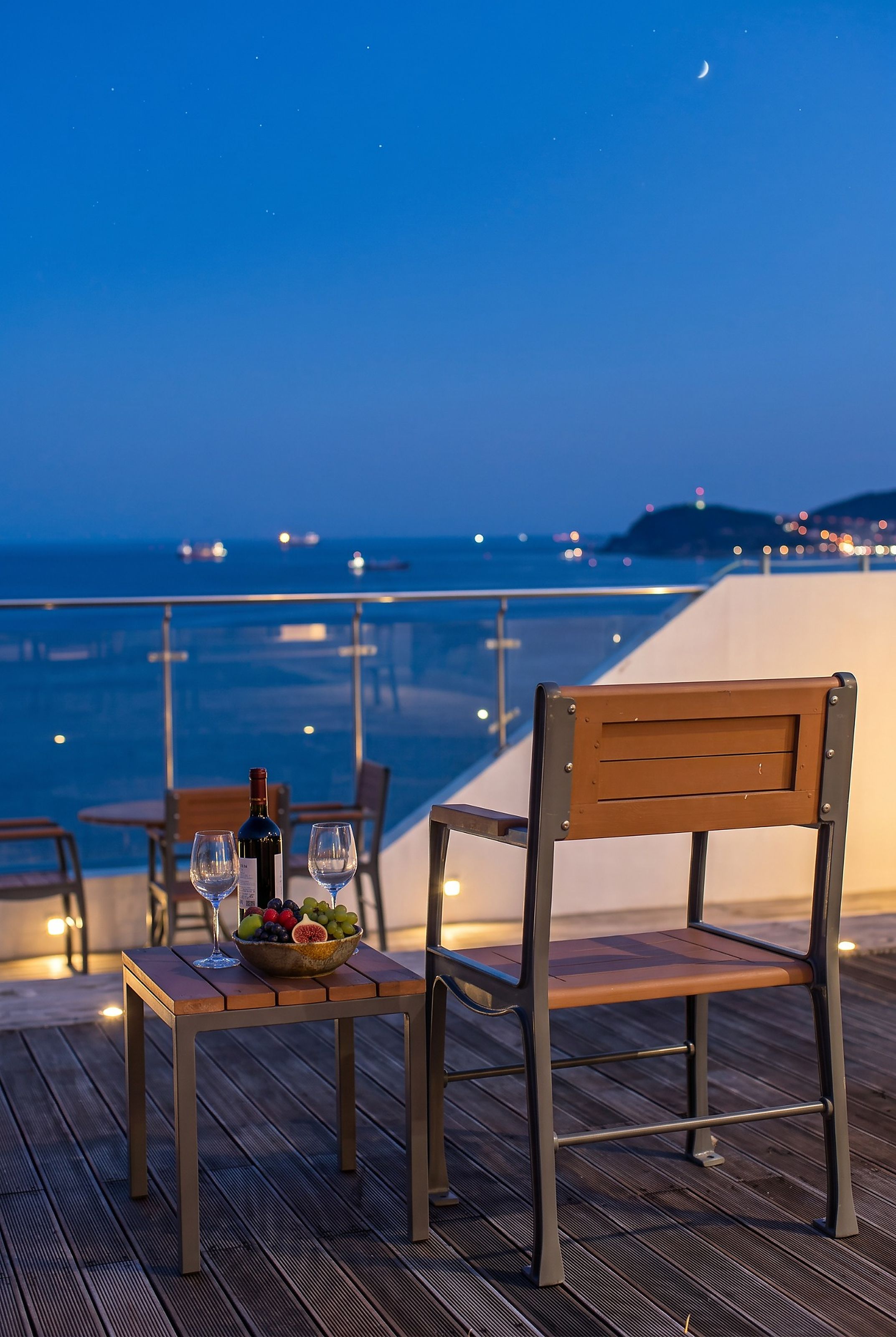 Une table avec une bouteille et des verres, un saladier de fruits et une chaise sur une terrasse en bois donnant sur la mer, le tout éclairé par la nuit.