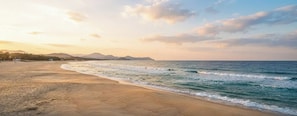 Una playa con una amplia orilla arenosa y olas suaves, con montañas al fondo.