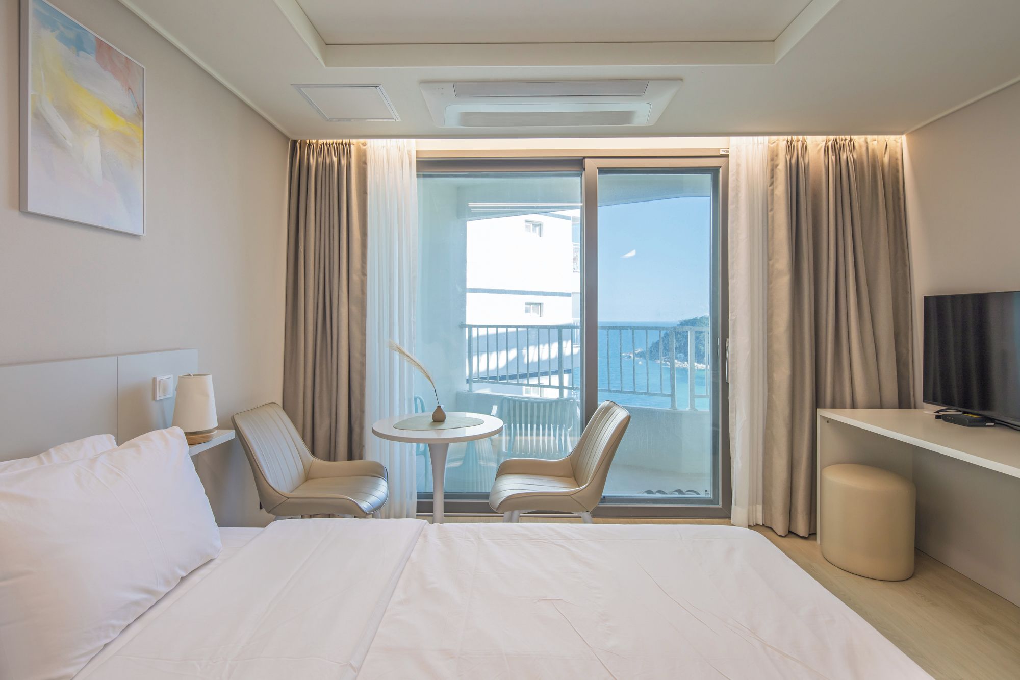 Ein Hotelzimmer mit einem Bett, zwei Sessel, einem kleinen Tisch, einem Fernseher und einem Balkon mit Vorhängen.