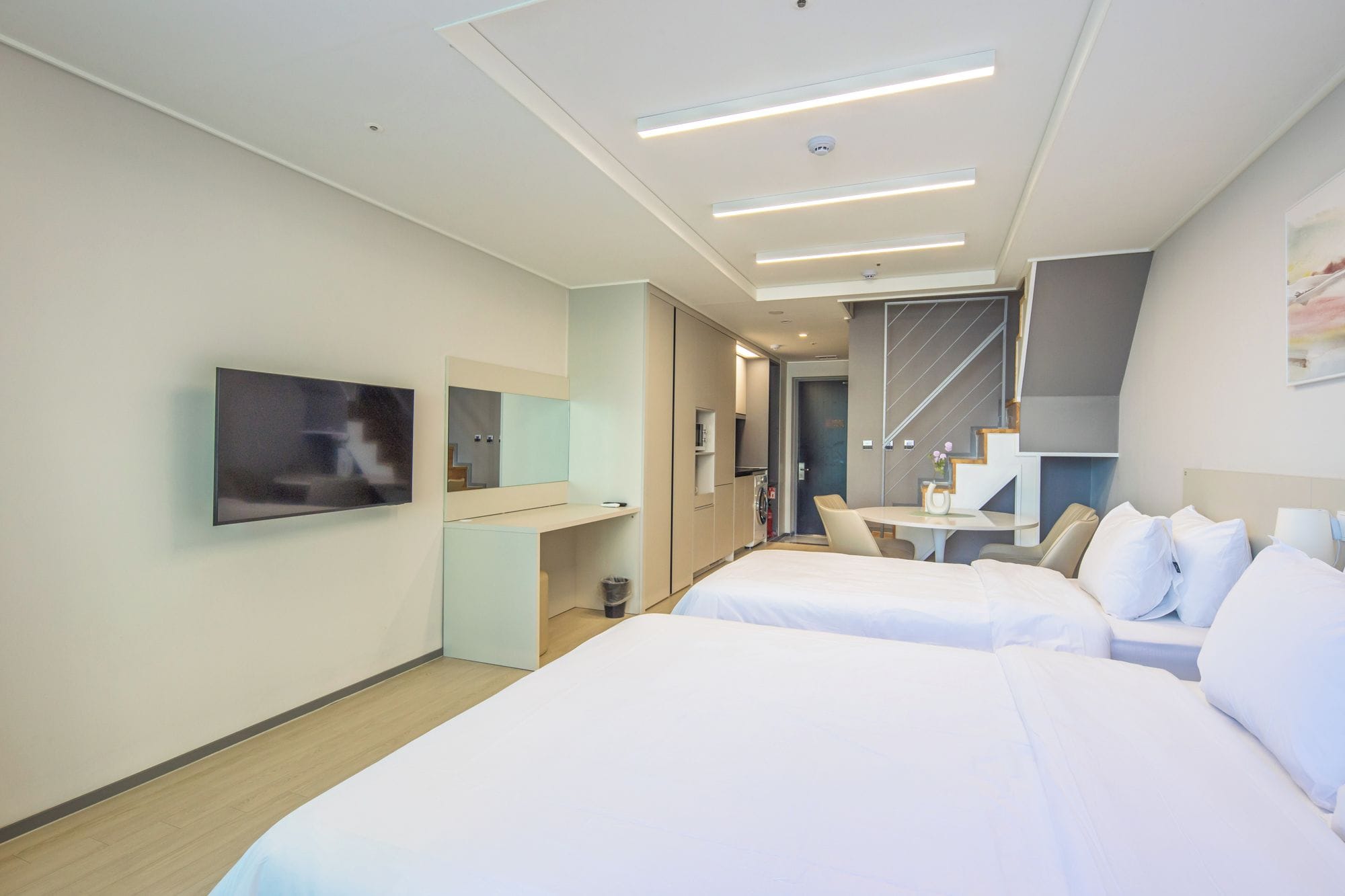 A modern hotel room with two beds, a flat-screen TV, a small kitchenette, and a dining area.