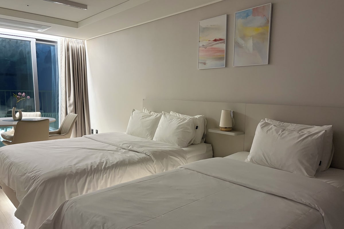 Habitación de hotel con dos camas, una mesita de noche, una lámpara y un ventanal con cortinas.